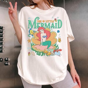 May include: Cream-coloured t-shirt featuring a graphic of Ariel from "The Little Mermaid." The design includes Ariel in a clam shell, with the words "The Little Mermaid" in green. Other characters from the movie are also featured.