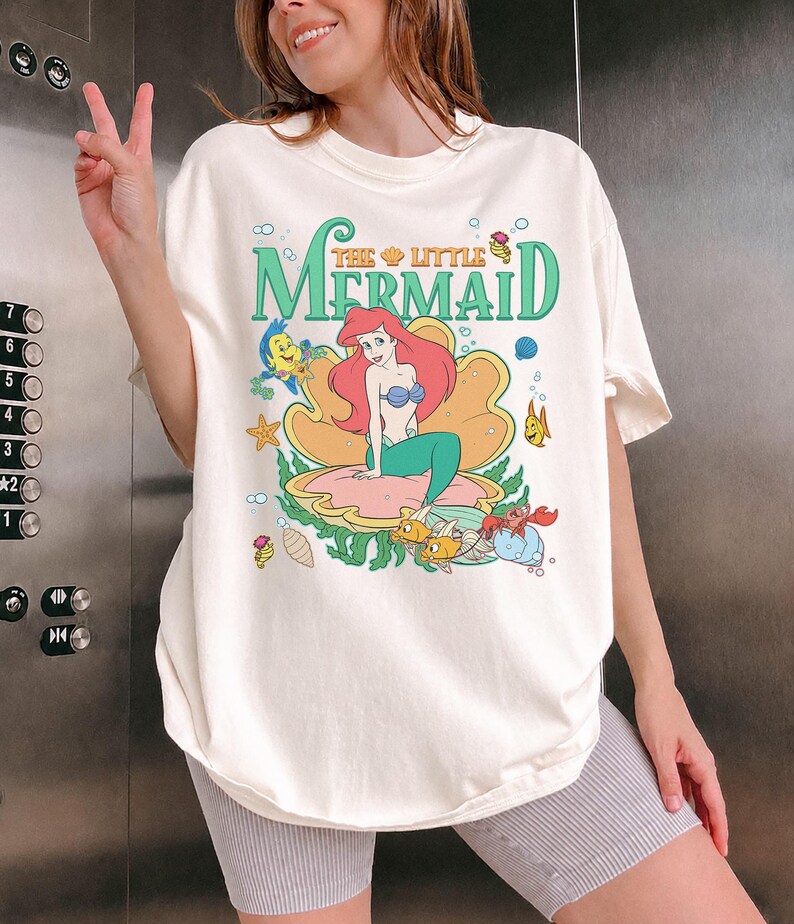 May include: Cream-coloured t-shirt featuring a graphic of Ariel from "The Little Mermaid." The design includes Ariel in a clam shell, with the words "The Little Mermaid" in green. Other characters from the movie are also featured.