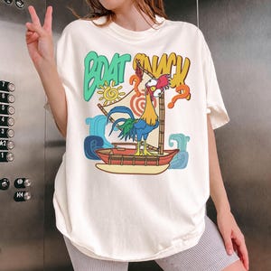 Puede incluir: Camiseta color crema con un gallo de dibujos animados navegando en un barco. La camiseta tiene las palabras "BOAT SNACK" en verde y amarillo, con un sol. El gallo es azul, amarillo y rojo, con un signo de interrogaci&oacute;n.