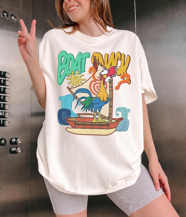 Puede incluir: Camiseta color crema con un gallo de dibujos animados navegando en un barco. La camiseta tiene las palabras "BOAT SNACK" en verde y amarillo, con un sol. El gallo es azul, amarillo y rojo, con un signo de interrogaci&oacute;n.