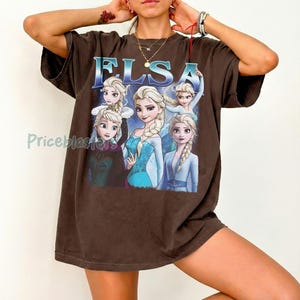 May include: Brown t-shirt featuring the name "ELSA" in blue letters above a graphic of the character Elsa from the movie Frozen. The graphic shows Elsa in various poses, wearing a blue dress and a white dress.