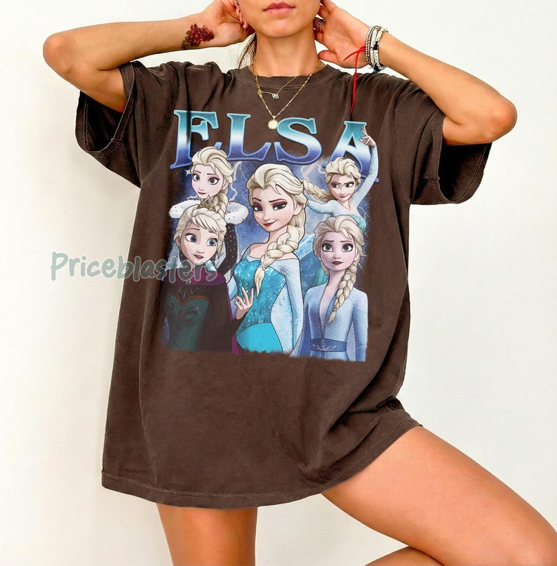 May include: Brown t-shirt featuring the name "ELSA" in blue letters above a graphic of the character Elsa from the movie Frozen. The graphic shows Elsa in various poses, wearing a blue dress and a white dress.