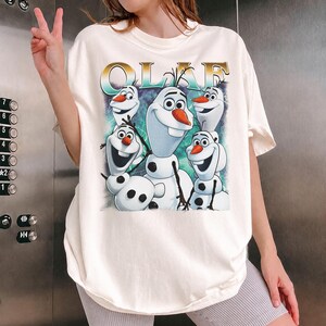 Puede incluir: Camiseta color crema con un gr&aacute;fico de Olaf de la pel&iacute;cula Frozen. La palabra "OLAF" est&aacute; impresa encima del personaje. Olaf se representa en varias poses con un fondo azul y verde.