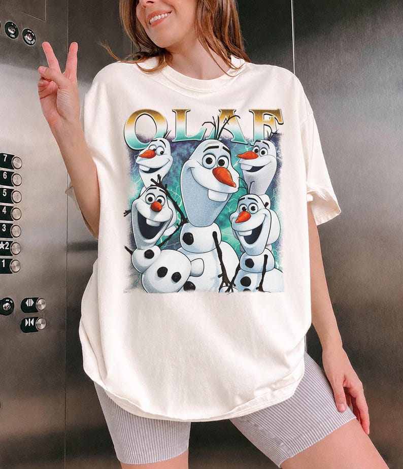 Puede incluir: Camiseta color crema con un gr&aacute;fico de Olaf de la pel&iacute;cula Frozen. La palabra "OLAF" est&aacute; impresa encima del personaje. Olaf se representa en varias poses con un fondo azul y verde.