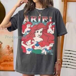 Pu&ograve; includere: T-shirt grigio scuro con una grafica di Ariel de La Sirenetta. Il design presenta Ariel con capelli rossi, un top blu e una coda verde, con il nome "ARIEL" in un font colorato sopra. La maglietta ha un aspetto vintage e usurato.