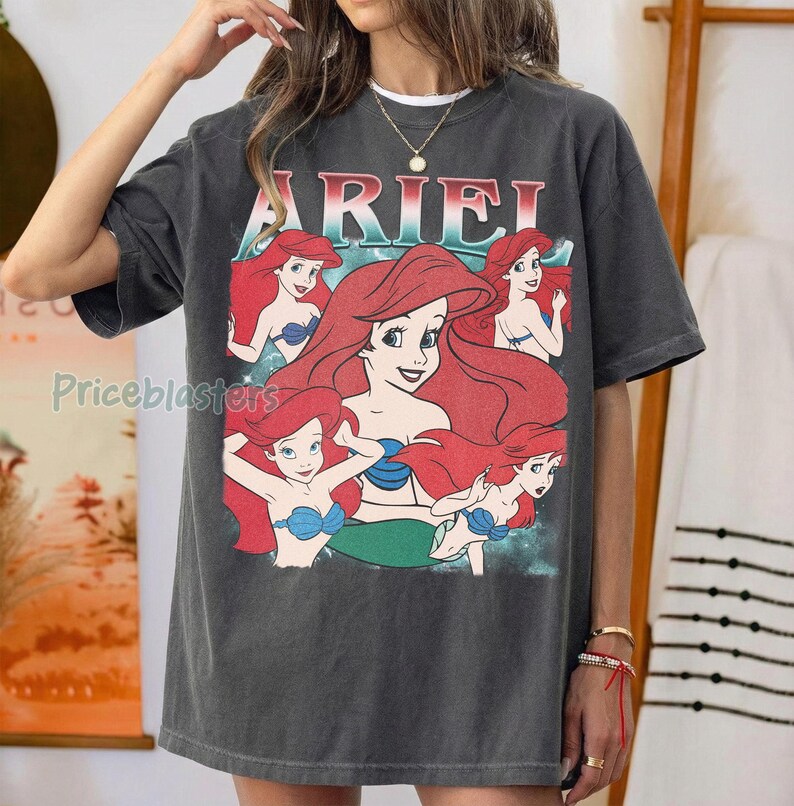 Pu&ograve; includere: T-shirt grigio scuro con una grafica di Ariel de La Sirenetta. Il design presenta Ariel con capelli rossi, un top blu e una coda verde, con il nome "ARIEL" in un font colorato sopra. La maglietta ha un aspetto vintage e usurato.
