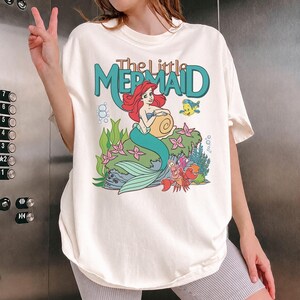May include: Cream-colored t-shirt featuring a graphic from "The Little Mermaid." The design includes Ariel, Flounder, and Sebastian, with the title in teal. The shirt has short sleeves and a relaxed fit.