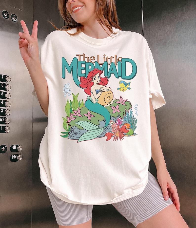 May include: Cream-colored t-shirt featuring a graphic from "The Little Mermaid." The design includes Ariel, Flounder, and Sebastian, with the title in teal. The shirt has short sleeves and a relaxed fit.
