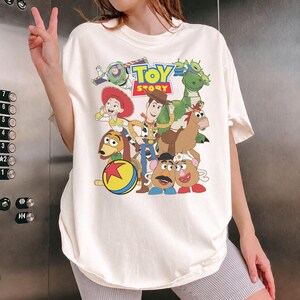 May include: Off-white t-shirt featuring a colorful graphic of characters from the animated film "Toy Story." The design includes Woody, Buzz Lightyear, Jessie, and other characters. The words "Toy Story" are printed in a playful font.