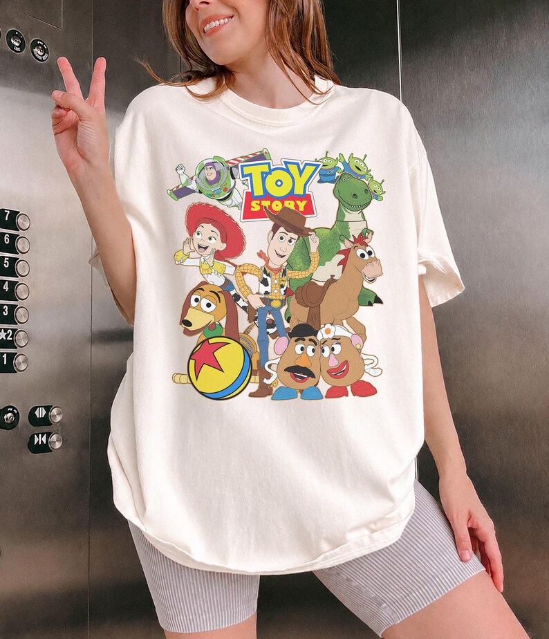 May include: Off-white t-shirt featuring a colorful graphic of characters from the animated film "Toy Story." The design includes Woody, Buzz Lightyear, Jessie, and other characters. The words "Toy Story" are printed in a playful font.