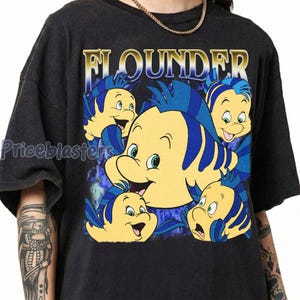 May include: Dark gray t-shirt featuring a graphic of Flounder from The Little Mermaid. The design includes multiple yellow and blue fish with the word "FLOUNDER" in gold lettering. The shirt has a vintage, distressed look.