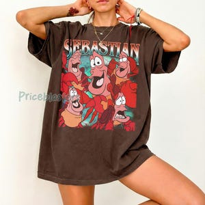 Pu&ograve; includere: T-shirt marrone con una grafica di Sebastian il granchio e altri personaggi de La Sirenetta. La parola "Sebastian" &egrave; stampata in alto con un carattere colorato. La maglietta ha maniche corte e una vestibilit&agrave; ampia.