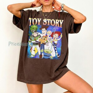 Puede incluir: Una camiseta marr&oacute;n con la inscripci&oacute;n "TOY STORY" en letras amarillas y naranjas. El gr&aacute;fico representa a los personajes de la pel&iacute;cula de animaci&oacute;n, incluyendo a Woody, Buzz Lightyear y Rex, sobre un fondo azul y morado.