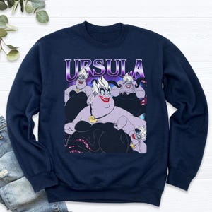 May include: Navy blue sweatshirt featuring a graphic of Ursula from The Little Mermaid. The design includes the word "URSULA" in a purple gradient font above a cartoon illustration of Ursula in various poses.