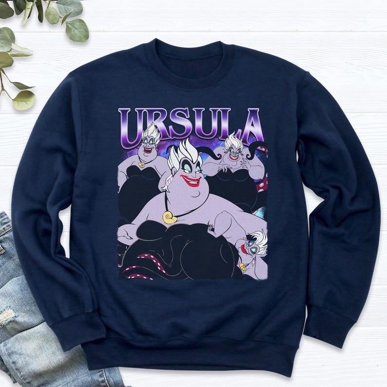 May include: Navy blue sweatshirt featuring a graphic of Ursula from The Little Mermaid. The design includes the word "URSULA" in a purple gradient font above a cartoon illustration of Ursula in various poses.