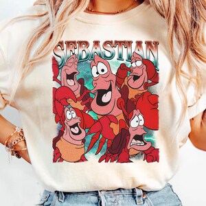 Pu&ograve; includere: T-shirt color crema con una grafica colorata del personaggio Sebastian de La Sirenetta. La parola "SEBASTIAN" &egrave; stampata sopra l'immagine in un font retr&ograve;. La grafica mostra diversi granchi con espressioni facciali.