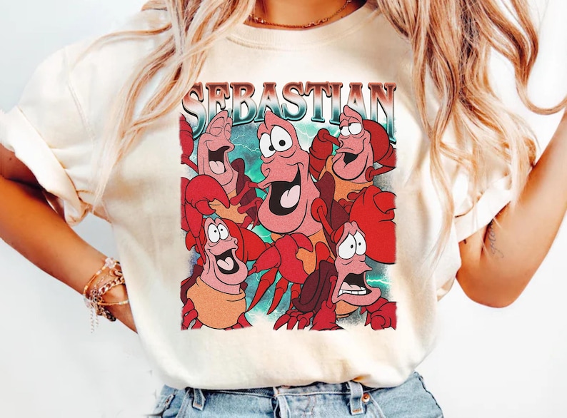 Pu&ograve; includere: T-shirt color crema con una grafica colorata del personaggio Sebastian de La Sirenetta. La parola "SEBASTIAN" &egrave; stampata sopra l'immagine in un font retr&ograve;. La grafica mostra diversi granchi con espressioni facciali.