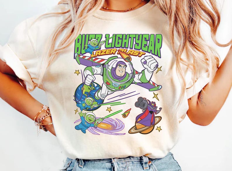 May include: Cream-colored t-shirt featuring a colorful graphic of Buzz Lightyear with the text "Buzz Lightyear Lazer Blast." The design includes cartoon characters, planets, and stars, creating a playful and dynamic composition.