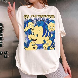 May include: Cream-colored t-shirt featuring a graphic of the character Flounder from The Little Mermaid. The design includes multiple yellow and blue fish with the word "FLOUNDER" in gold lettering. The shirt has short sleeves and a relaxed fit.