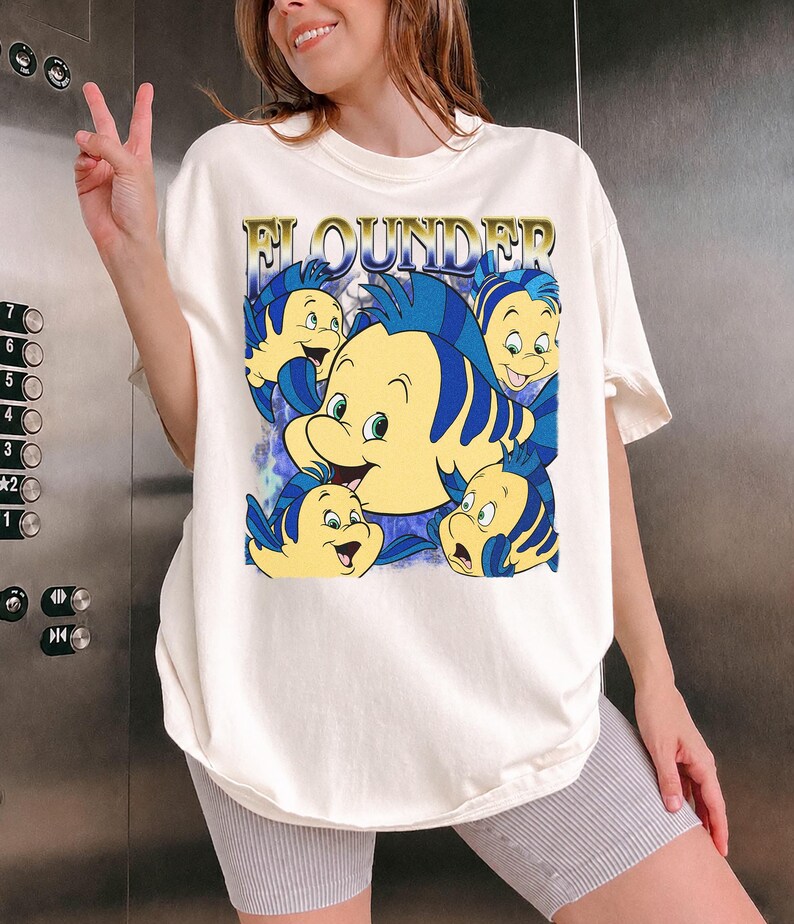 May include: Cream-colored t-shirt featuring a graphic of the character Flounder from The Little Mermaid. The design includes multiple yellow and blue fish with the word "FLOUNDER" in gold lettering. The shirt has short sleeves and a relaxed fit.