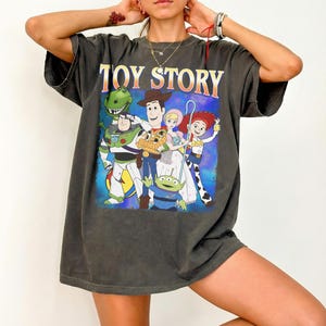Puede incluir: Camiseta gris oscuro con las palabras "TOY STORY" en naranja y amarillo sobre un gr&aacute;fico de personajes de la pel&iacute;cula animada. El gr&aacute;fico incluye a Buzz Lightyear, Woody y otros personajes sobre un fondo azul.