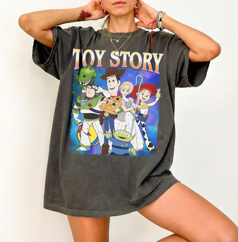 Puede incluir: Camiseta gris oscuro con las palabras "TOY STORY" en naranja y amarillo sobre un gr&aacute;fico de personajes de la pel&iacute;cula animada. El gr&aacute;fico incluye a Buzz Lightyear, Woody y otros personajes sobre un fondo azul.
