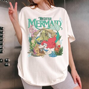 Może przedstawiać: Kremowy t-shirt z grafiką z filmu "Mała Syrenka". Projekt zawiera Ariel, Florka, Sebastiana i zamek, z napisem "The Little Mermaid" w kolorze zielonym. Koszulka ma luźny kr&oacute;j.