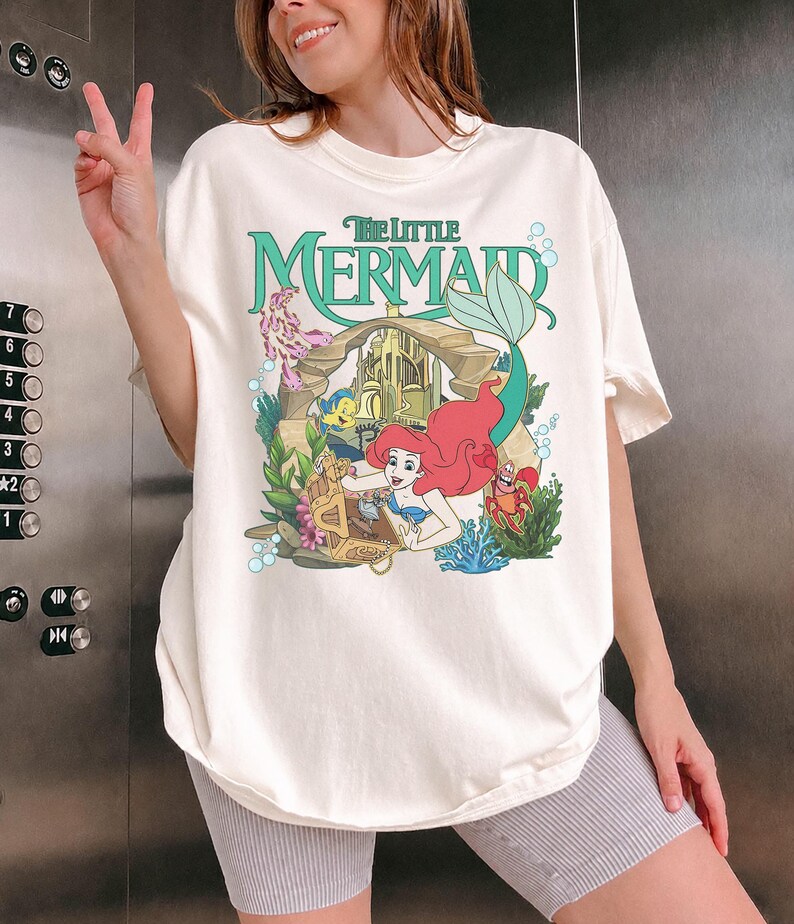 Może przedstawiać: Kremowy t-shirt z grafiką z filmu "Mała Syrenka". Projekt zawiera Ariel, Florka, Sebastiana i zamek, z napisem "The Little Mermaid" w kolorze zielonym. Koszulka ma luźny kr&oacute;j.