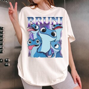 May include: Cream-colored t-shirt featuring a graphic of Bruni, a blue salamander character, with the word "BRUNI" in a purple and blue font. The salamanders have large eyes and are set against a purple lightning background.