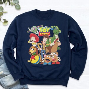 May include: Navy blue sweatshirt featuring a colorful graphic of characters from the animated film "Toy Story." The design includes Woody, Buzz Lightyear, Jessie, and other beloved characters. The text "Toy Story" is also displayed.