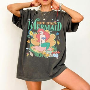 May include: Dark grey t-shirt with a colourful graphic of Ariel from The Little Mermaid. The design includes Ariel, Flounder, Sebastian, and the text "THE LITTLE MERMAID" in green and blue. The shirt has a vintage, worn appearance.