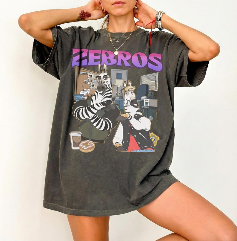 Pu&ograve; includere: T-shirt grigio scuro con un'illustrazione a cartoni animati di due zebre in un ufficio. La parola "ZEBROS" &egrave; stampata in alto in una sfumatura viola e rosa. Una zebra indossa una giacca e l'altra un abito.