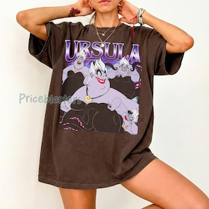 May include: Brown t-shirt featuring a graphic of Ursula from The Little Mermaid, with the name "URSULA" in purple. The design includes Ursula's face and body, with her tentacles visible. The shirt is a casual, oversized style.