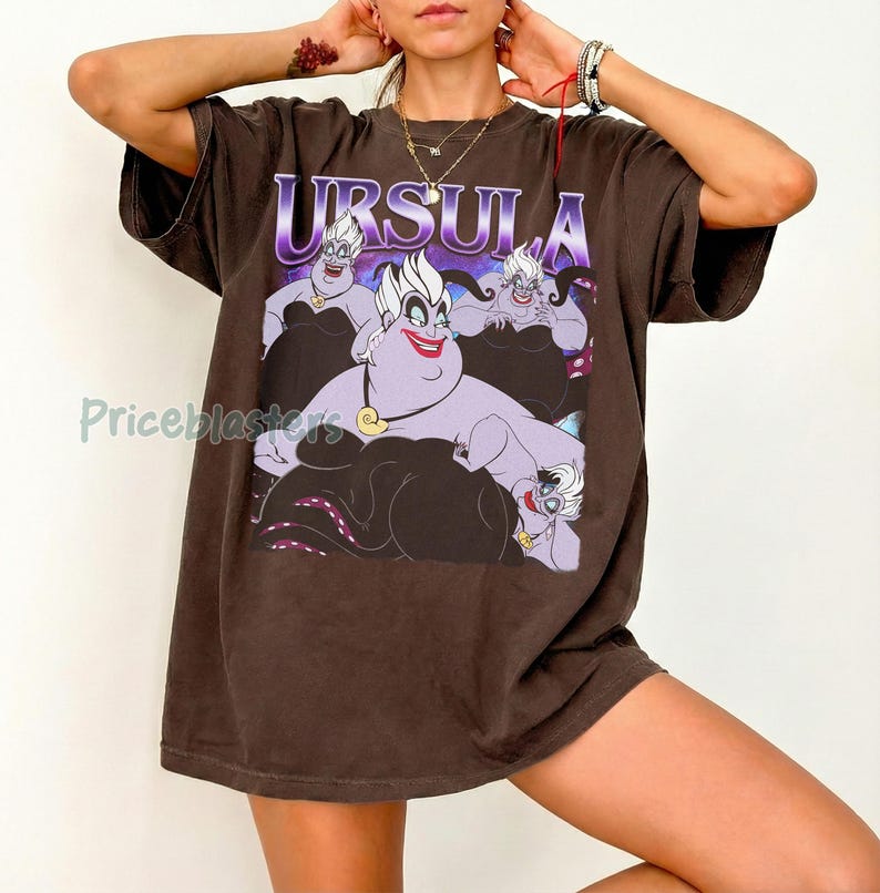 May include: Brown t-shirt featuring a graphic of Ursula from The Little Mermaid, with the name "URSULA" in purple. The design includes Ursula's face and body, with her tentacles visible. The shirt is a casual, oversized style.