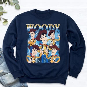 May include: Navy blue sweatshirt featuring a graphic of Woody from Toy Story. The design includes multiple images of Woody with a blue lightning background and the word "WOODY" in gold lettering.