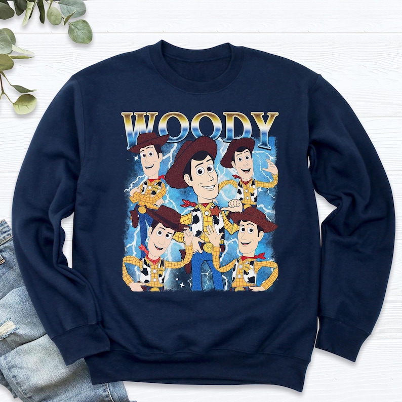 May include: Navy blue sweatshirt featuring a graphic of Woody from Toy Story. The design includes multiple images of Woody with a blue lightning background and the word "WOODY" in gold lettering.