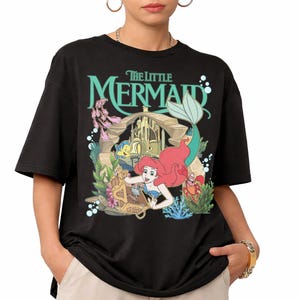 Może przedstawiać: Czarny t-shirt z kolorową grafiką z filmu "Mała Syrenka". Projekt zawiera Ariel, Florka, Sebastiana i skrzynię skarb&oacute;w. Tytuł "The Little Mermaid" jest w turkusowych literach nad sceną.