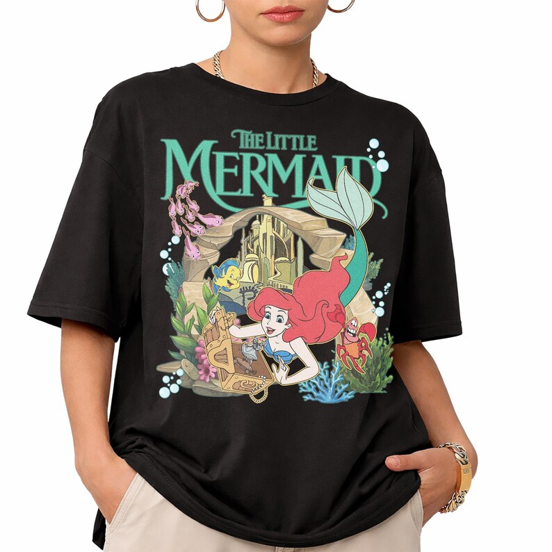 Może przedstawiać: Czarny t-shirt z kolorową grafiką z filmu "Mała Syrenka". Projekt zawiera Ariel, Florka, Sebastiana i skrzynię skarb&oacute;w. Tytuł "The Little Mermaid" jest w turkusowych literach nad sceną.