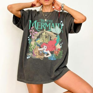Może przedstawiać: Ciemnoszara koszulka z grafiką z filmu "Mała Syrenka". Projekt zawiera Ariel, Florka, Sebastiana i scenę zamku. Tekst "The Little Mermaid" jest w kolorze morskim. Koszulka ma kr&oacute;tkie rękawy i luźny kr&oacute;j.