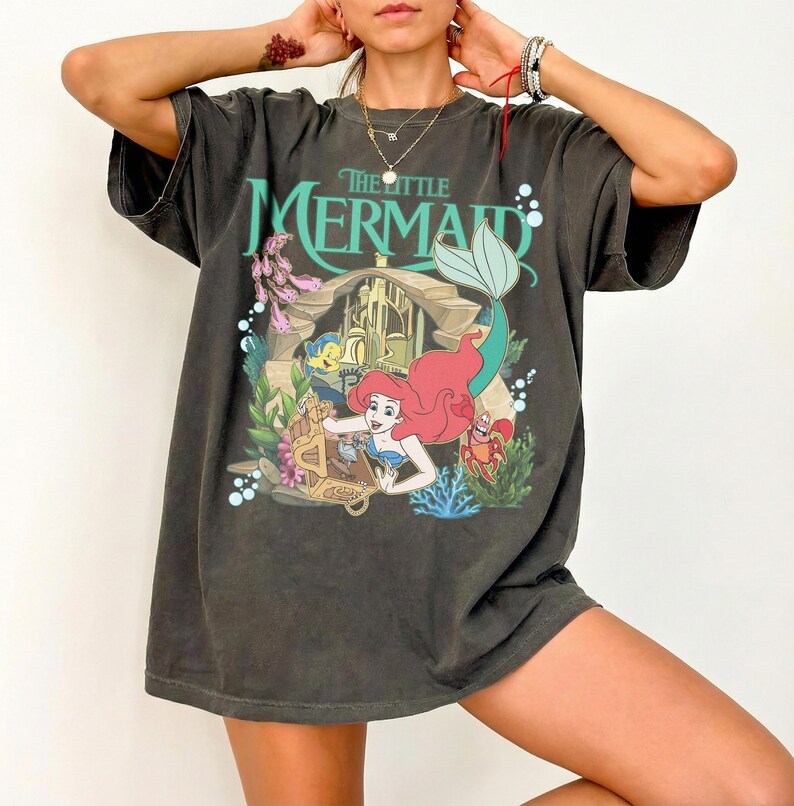 Może przedstawiać: Ciemnoszara koszulka z grafiką z filmu "Mała Syrenka". Projekt zawiera Ariel, Florka, Sebastiana i scenę zamku. Tekst "The Little Mermaid" jest w kolorze morskim. Koszulka ma kr&oacute;tkie rękawy i luźny kr&oacute;j.