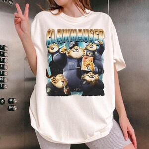 Puede incluir: Camiseta color crema con un gr&aacute;fico de Clawhauser de Zootopia. El dise&ntilde;o incluye a Clawhauser con uniforme de polic&iacute;a, con el texto "CLAWHAUSER" en fuente azul y dorada. La camiseta es de corte holgado.