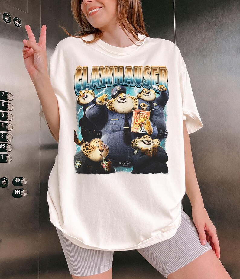 Puede incluir: Camiseta color crema con un gr&aacute;fico de Clawhauser de Zootopia. El dise&ntilde;o incluye a Clawhauser con uniforme de polic&iacute;a, con el texto "CLAWHAUSER" en fuente azul y dorada. La camiseta es de corte holgado.