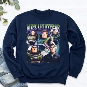 May include: Navy blue sweatshirt featuring a graphic of Buzz Lightyear and the text "BUZZ LIGHTYEAR" in green and purple. The graphic shows multiple images of Buzz Lightyear in various poses. The sweatshirt is made of a soft material.