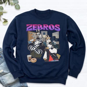 Pu&ograve; includere: Felpa blu navy con un'illustrazione a cartoni animati di due zebre in un ufficio. Le zebre hanno strisce bianche e nere, indossano giacche e tengono in mano del cibo. La parola "ZEBROS" &egrave; stampata in un gradiente viola e rosa.