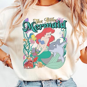 May include: Cream-colored t-shirt featuring a colorful graphic of Ariel from The Little Mermaid. The design includes Ariel with red hair, a dolphin, and other sea creatures, with the words "The Little Mermaid" in a decorative font.