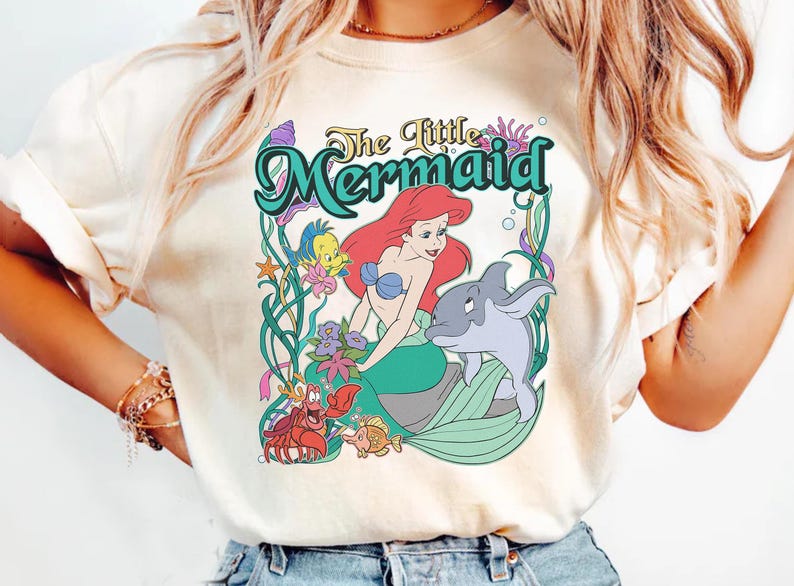 May include: Cream-colored t-shirt featuring a colorful graphic of Ariel from The Little Mermaid. The design includes Ariel with red hair, a dolphin, and other sea creatures, with the words "The Little Mermaid" in a decorative font.