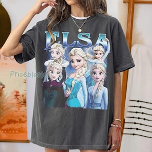 May include: Dark gray t-shirt featuring the word "ELSA" in blue, with multiple images of the character Elsa from the movie Frozen. The images show Elsa in various poses and outfits. The shirt is a casual, oversized style.