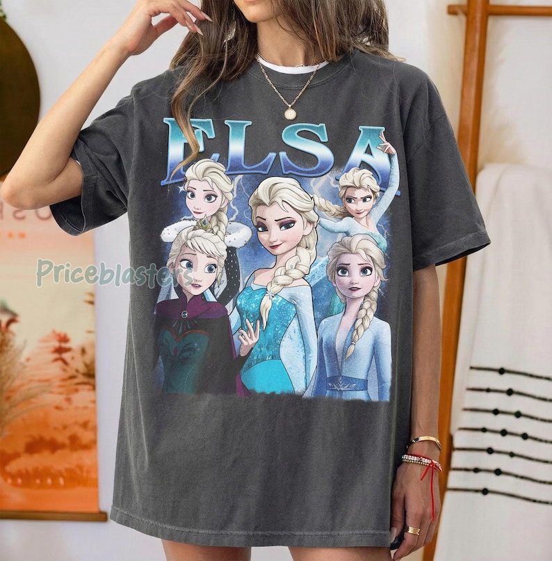 May include: Dark gray t-shirt featuring the word "ELSA" in blue, with multiple images of the character Elsa from the movie Frozen. The images show Elsa in various poses and outfits. The shirt is a casual, oversized style.