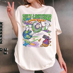 May include: Off-white t-shirt featuring a colorful graphic of Buzz Lightyear in a space scene with the text "Buzz Lightyear Laser Blast" in green and orange. The design includes cartoon aliens, planets, and stars.