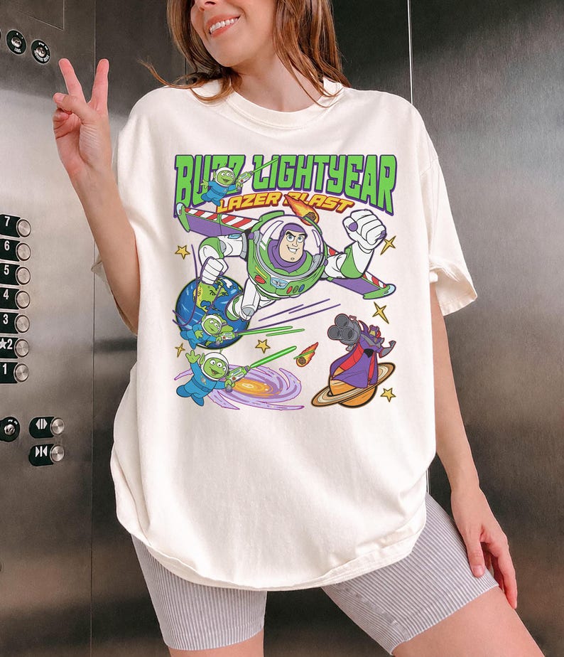 May include: Off-white t-shirt featuring a colorful graphic of Buzz Lightyear in a space scene with the text "Buzz Lightyear Laser Blast" in green and orange. The design includes cartoon aliens, planets, and stars.